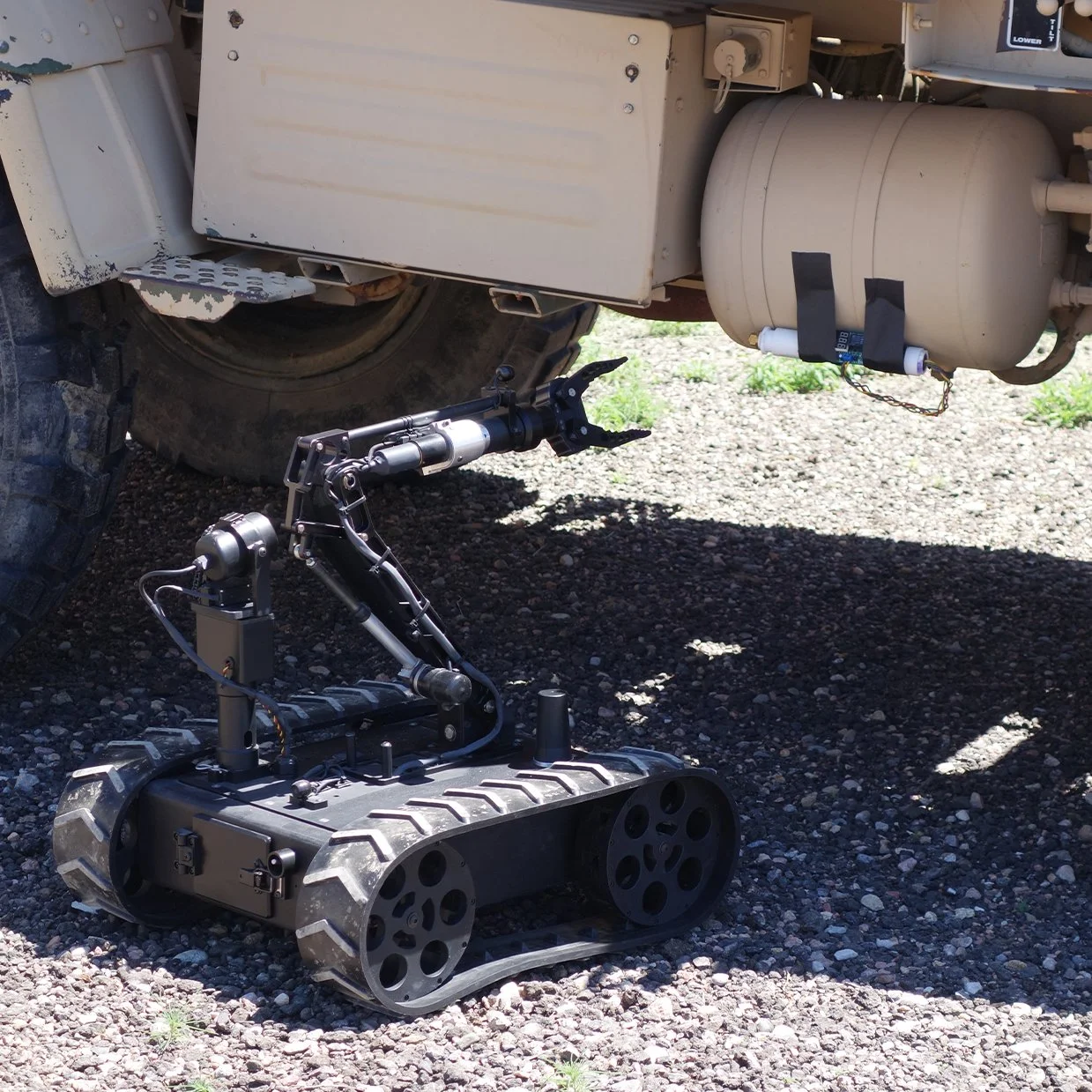 An MMP-30-EOD robot with tank-like treads and a robotic arm extends under a large industrial vehicle, which appears to be a military or construction vehicle, with gravel ground and some grass in the background.