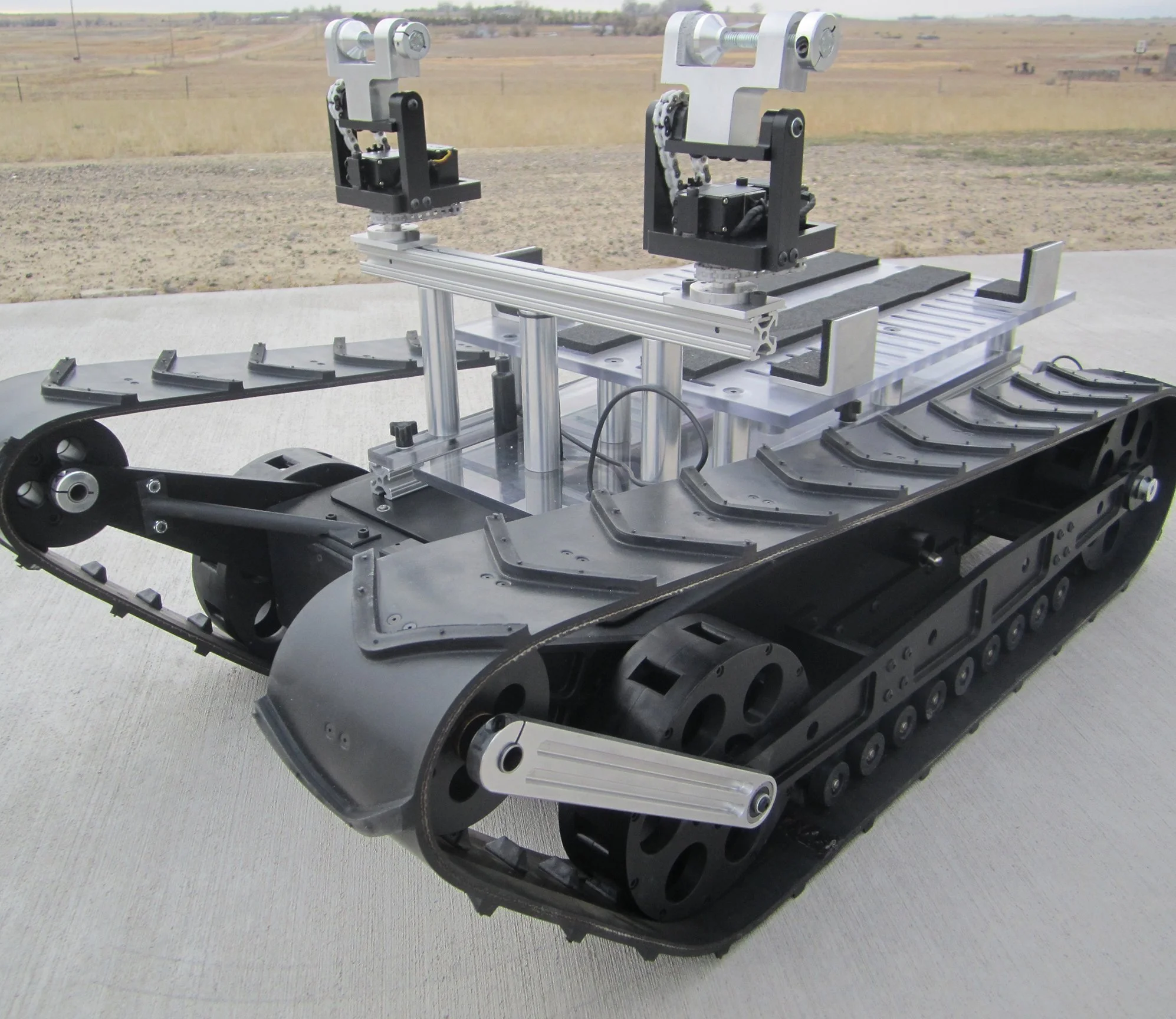 A prototype robotic platform with tank tracks and two robotic arms on a concrete surface outdoors, with open fields in the background.