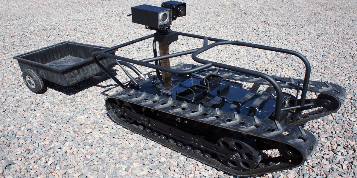 A small tracked robot with a black metal frame, mounted sensors or cameras, and a cart attached at the rear, situated on gravel ground.