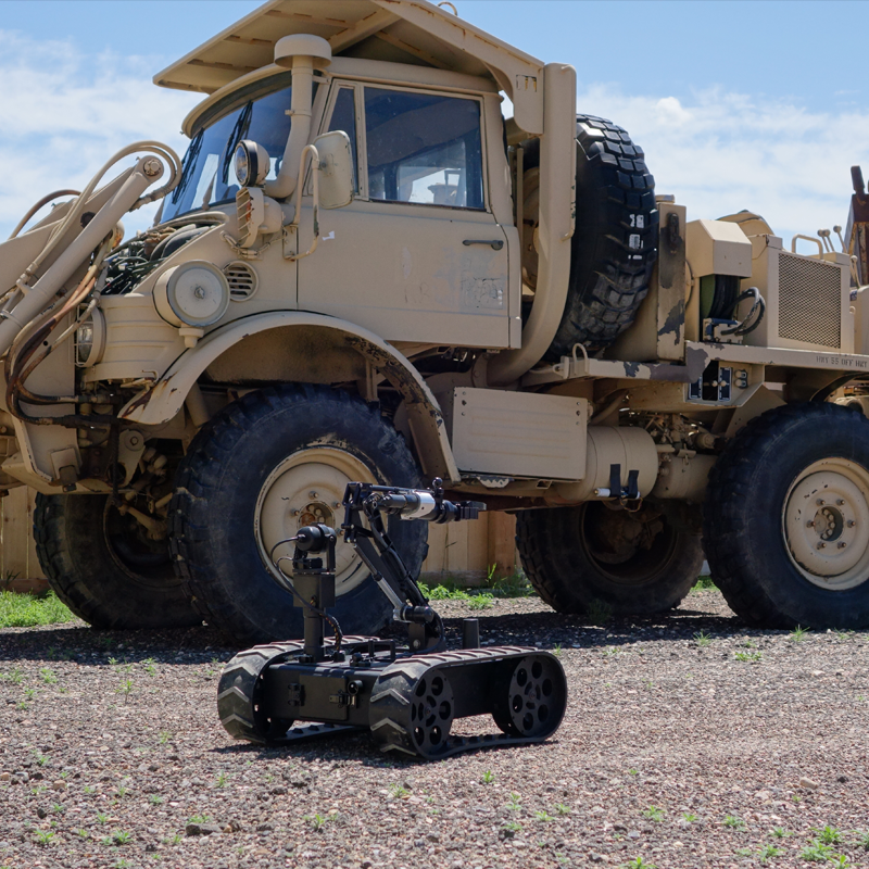A large beige military vehicle with oversized tires, parked outdoors against a wooden fence and blue sky. In front of the vehicle, a small robot with treads and a camera or sensor is positioned on the ground.