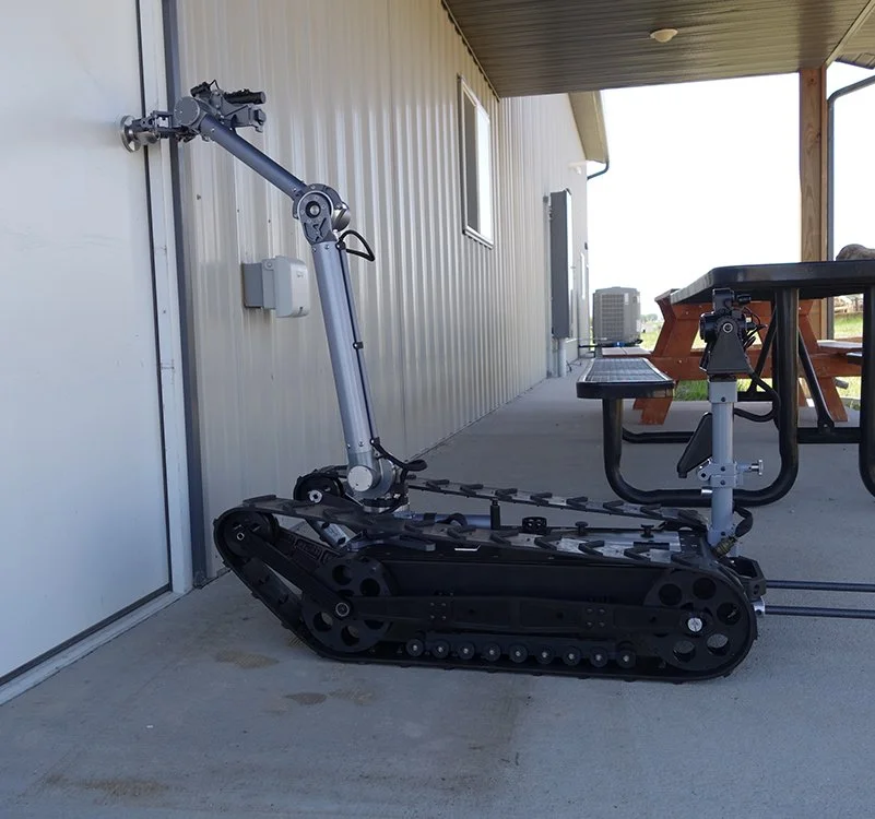 A robot with tank-like tracks and an articulated arm, positioned on a concrete porch beside a white building, with a picnic table and a bench in the background. MMP-40-EOD robot.