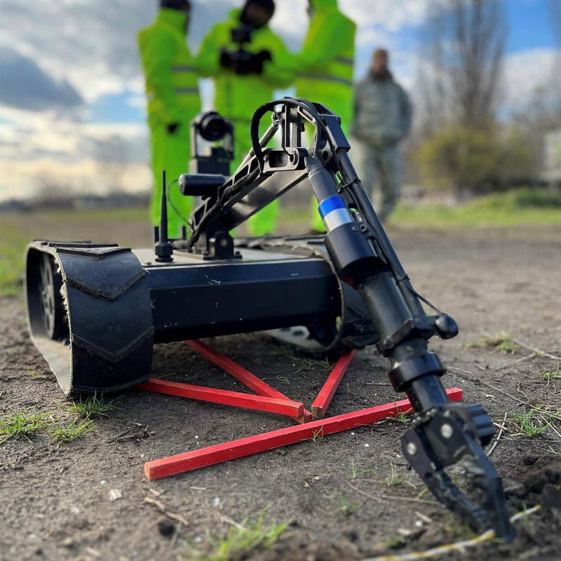 An EOD robot with tank-like tracks and a mechanical arm, positioned on the ground outdoors, with two people in reflective safety vests and another person in casual clothing observing in the background.