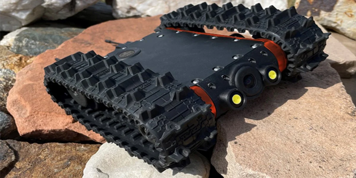 A compact robot with tank treads, black and orange body, and multiple camera lenses, placed on rocks outdoors.