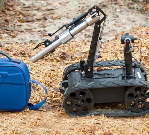 Small EOD robot with mechanical arm and mounted disruptor cannon, on a dirt ground with fallen leaves.