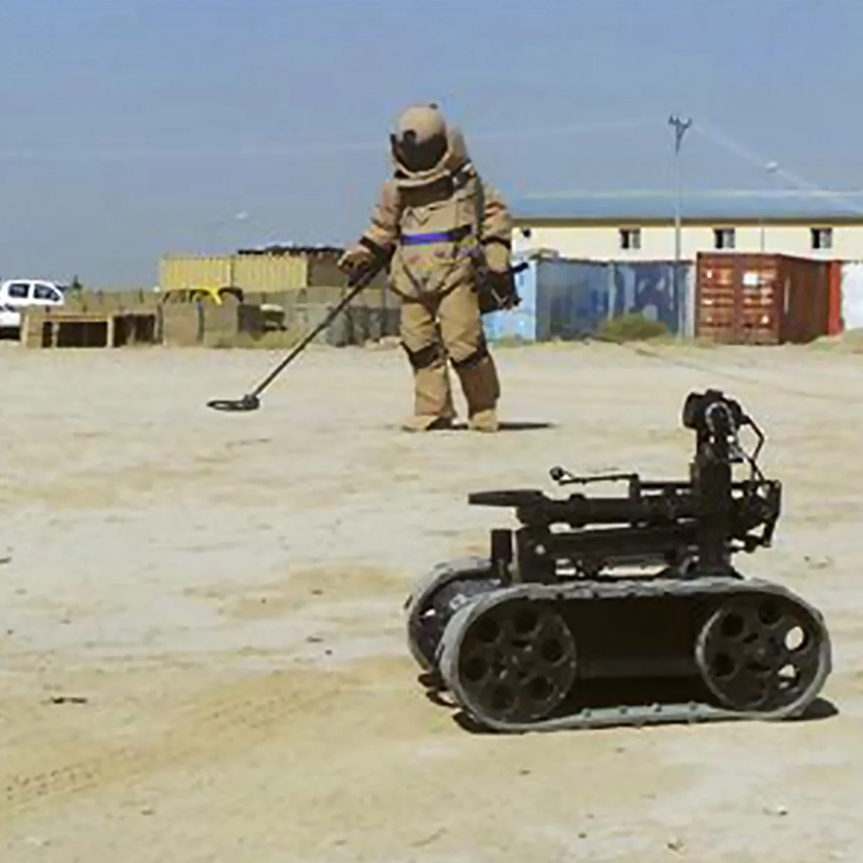 An EOD technician using a metal detector outdoors on a sandy surface, with an MMP-30-EOD robot nearby and some buildings and structures in the background.