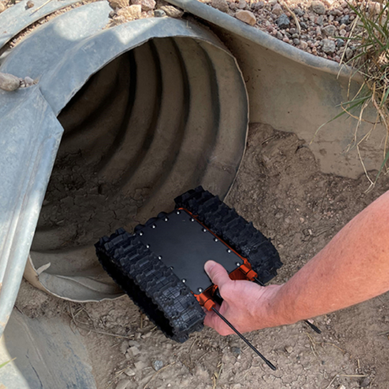 A person holding a MicroTraxx inspection robot with tank tracks, emerging from a large metal pipe in the ground, possibly for exploration or inspection.