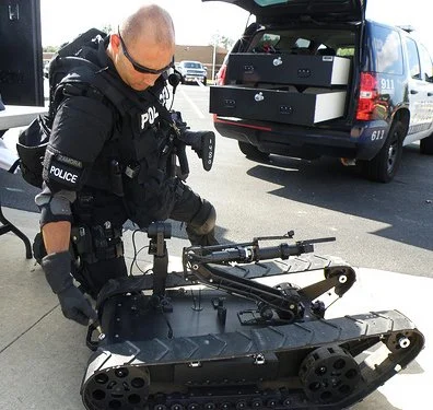 Police officer handling a robotic robot with tank-like tracks near a police vehicle.
