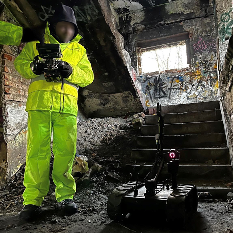 The MMP-30-EOD robot and operator in high-visibility yellow protective suit and gloves with blurred face operating a surveying drone inside a dilapidated, graffiti-covered building with broken stairs and a window.