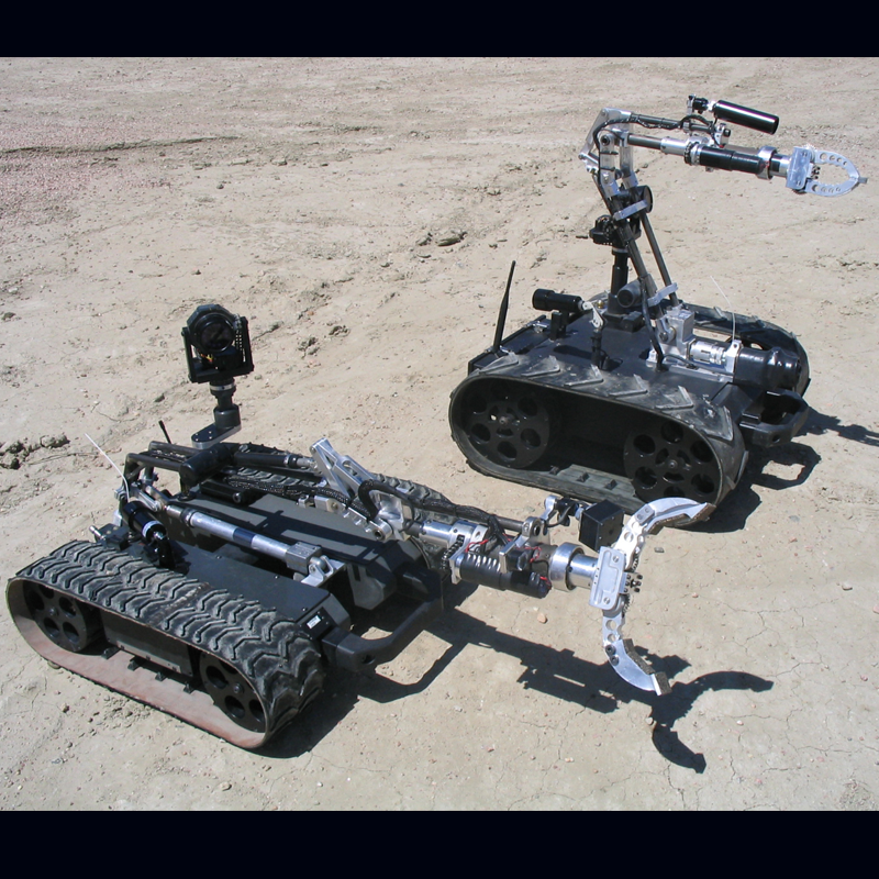 Two small robotic vehicles on sandy terrain with tracks and mechanical arms