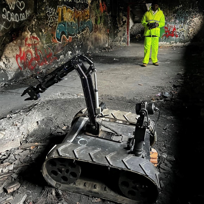 The MMP-30-EOD robot with a robotic arm, multiple cameras, in an underground tunnel with graffiti on the walls. A man in a high-visibility yellow suit stands in the background holding a remote control.