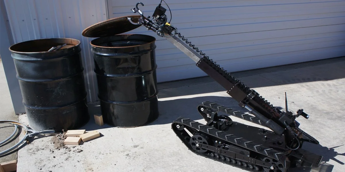 A small tracked robot with a mechanical arm extended upward, positioned outdoors next to two large black metal barrels and some bricks on a concrete surface.