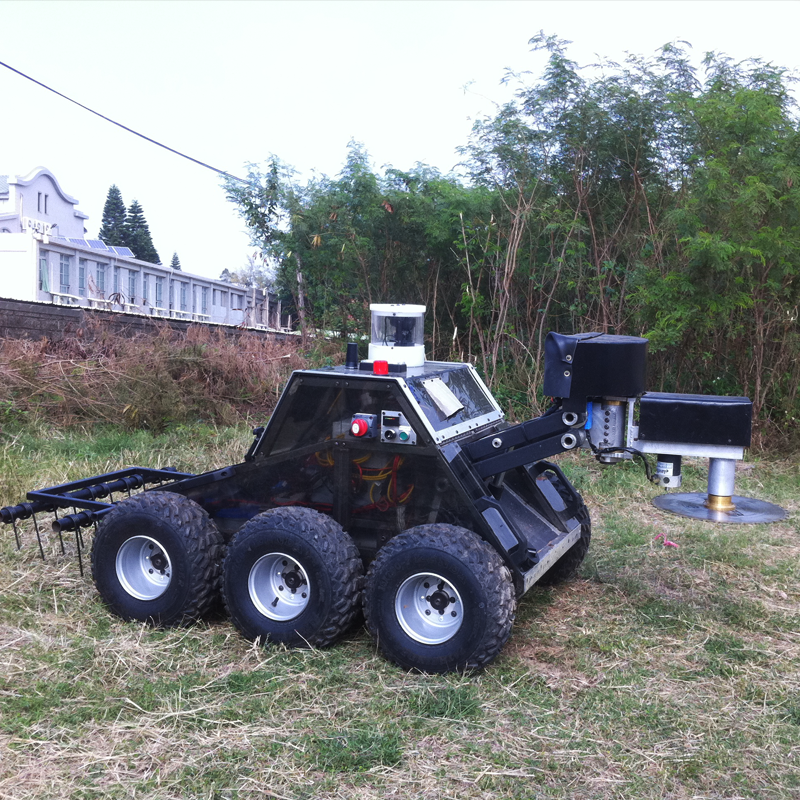 An EOD robot with a mechanical arm extending from its body, situated on a grassy field with trees and a building in the background used for clearing mine fields.