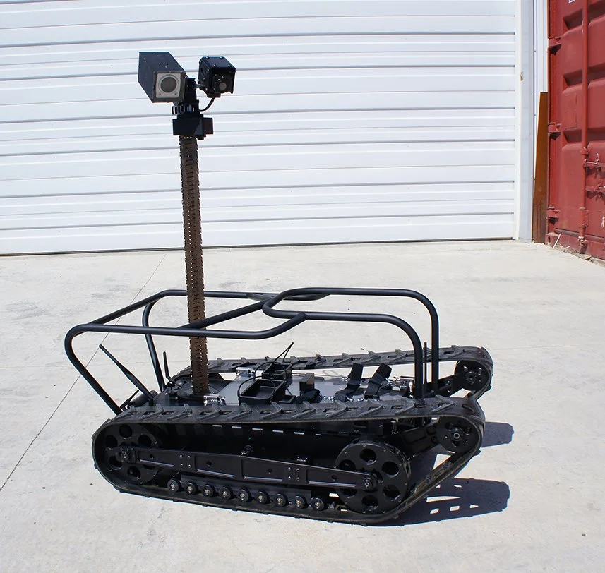 A small robotic vehicle with tank treads, a metal frame, and a camera mounted on a telescoping post, positioned outdoors in front of a white garage door. MMP-40X.