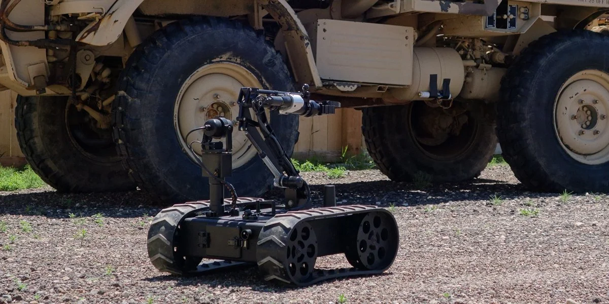 A small, robotic tracked vehicle with a mechanical arm, in front of a large military vehicle with oversized tires.