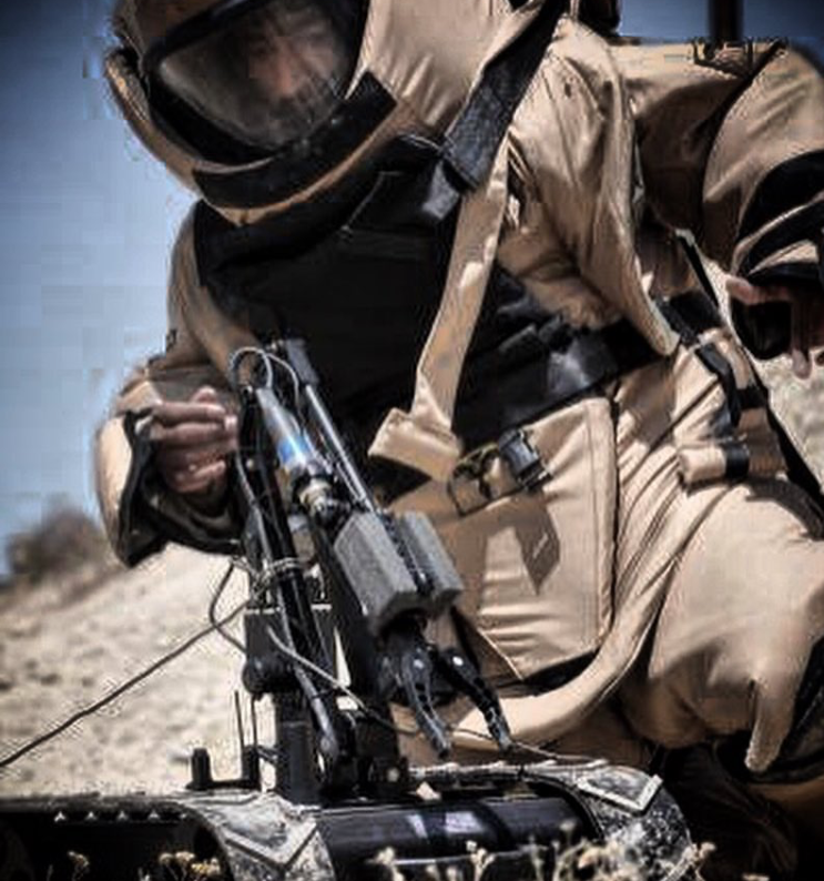 An EOD tech in desert camouflage military gear wearing a helmet with a visor, operating a drone or robotic device outdoors in a desert landscape.