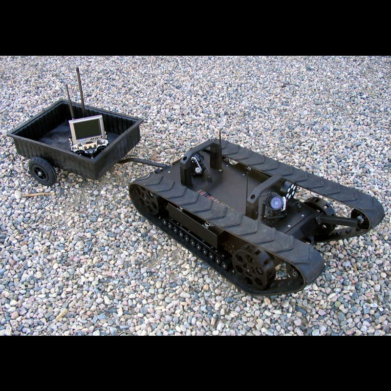 A remotely controlled robotic UGV with caterpillar tracks on gravel ground, connected to a black remote control box with antennas and a display.