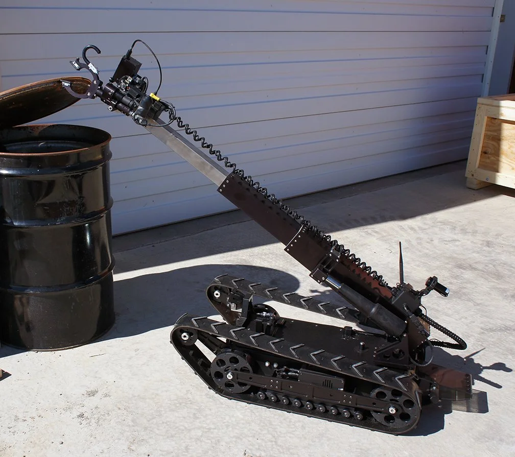 A robot resembling a small tank with a long mechanical arm and claw, positioned outdoors on a concrete surface near a black barrel and a white garage door.
