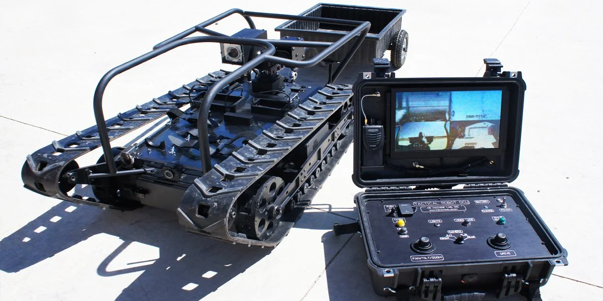 A remote-controlled military robot with treads, a camera, and a rugged control box with a screen and joysticks on concrete.