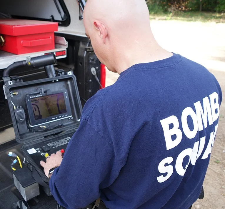 A person in a navy blue shirt with 'BOMB SQUAD' written on the back is operating a portable electronic device inside a black case, with a red toolbox and vehicle in the background.