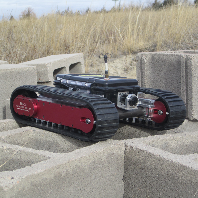 A robotic vehicle with Track Pods, a black body, and various mechanical components, situated outdoors among concrete blocks and dry grass.