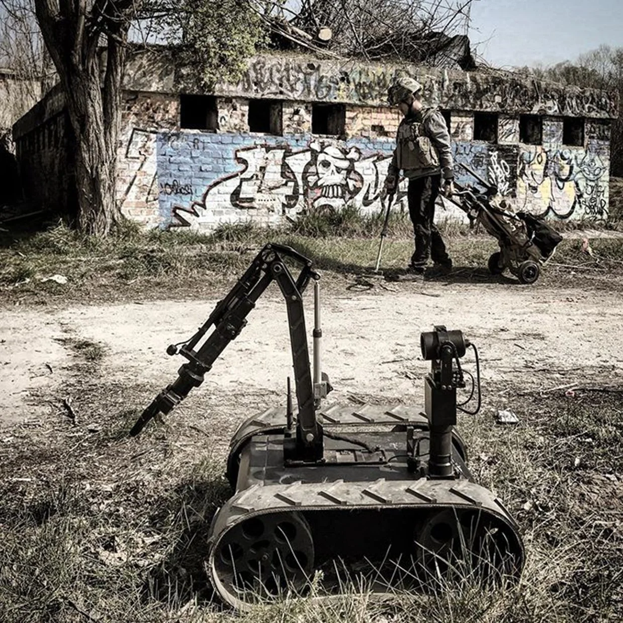 A person in military attire operating a small robotic vehicle with tracks on a dirt area in front of a graffiti-covered brick building. The graffiti features a skull and cartoon characters.