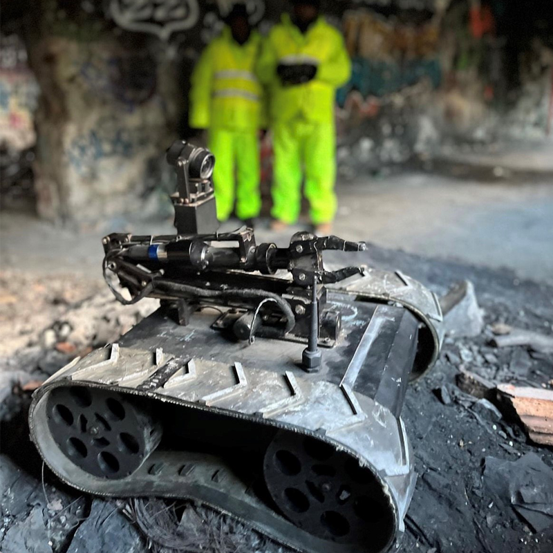 An MMP-30-EOD robot with caterpillar treads and mounted camera or sensor equipment on a destroyed surface, with two people in high-visibility yellow suits in the background.