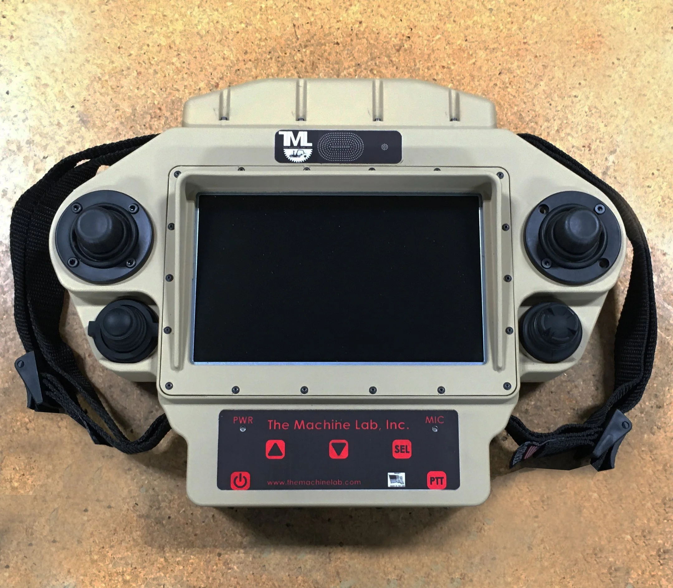 A handheld robot controller with a gray body and black buttons, labeled with the brand "The Machine Lab, Inc." and various controls, resting on a brown speckled surface.