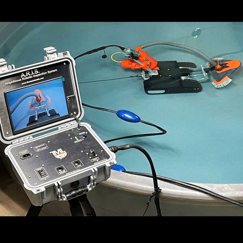 A robotic amphibious inspection device submerged in a pool connected to a control unit with a screen showing the device underwater.