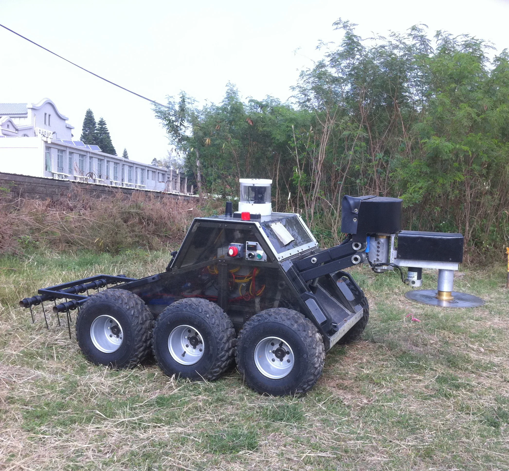 A small robot or vehicle with six wheels, a camera or sensor on top, and connected components, situated outdoors near trees and a building.