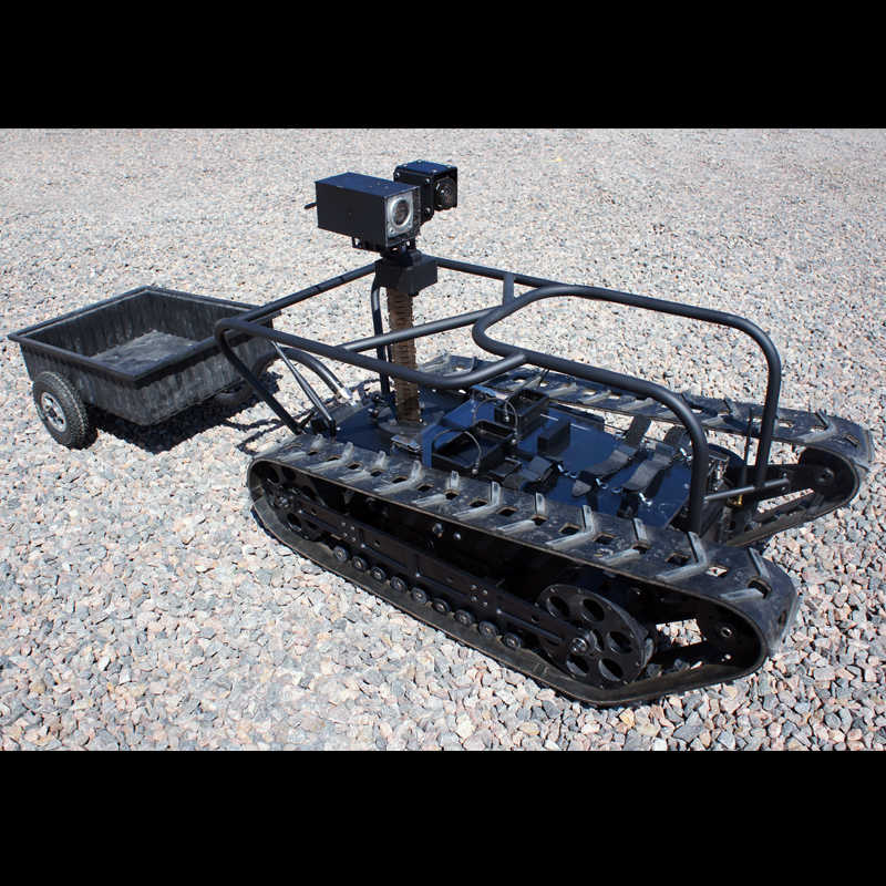 A robotic ground vehicle with tank-like tracks, mounted cameras, and a small trailer on a gravel surface.
