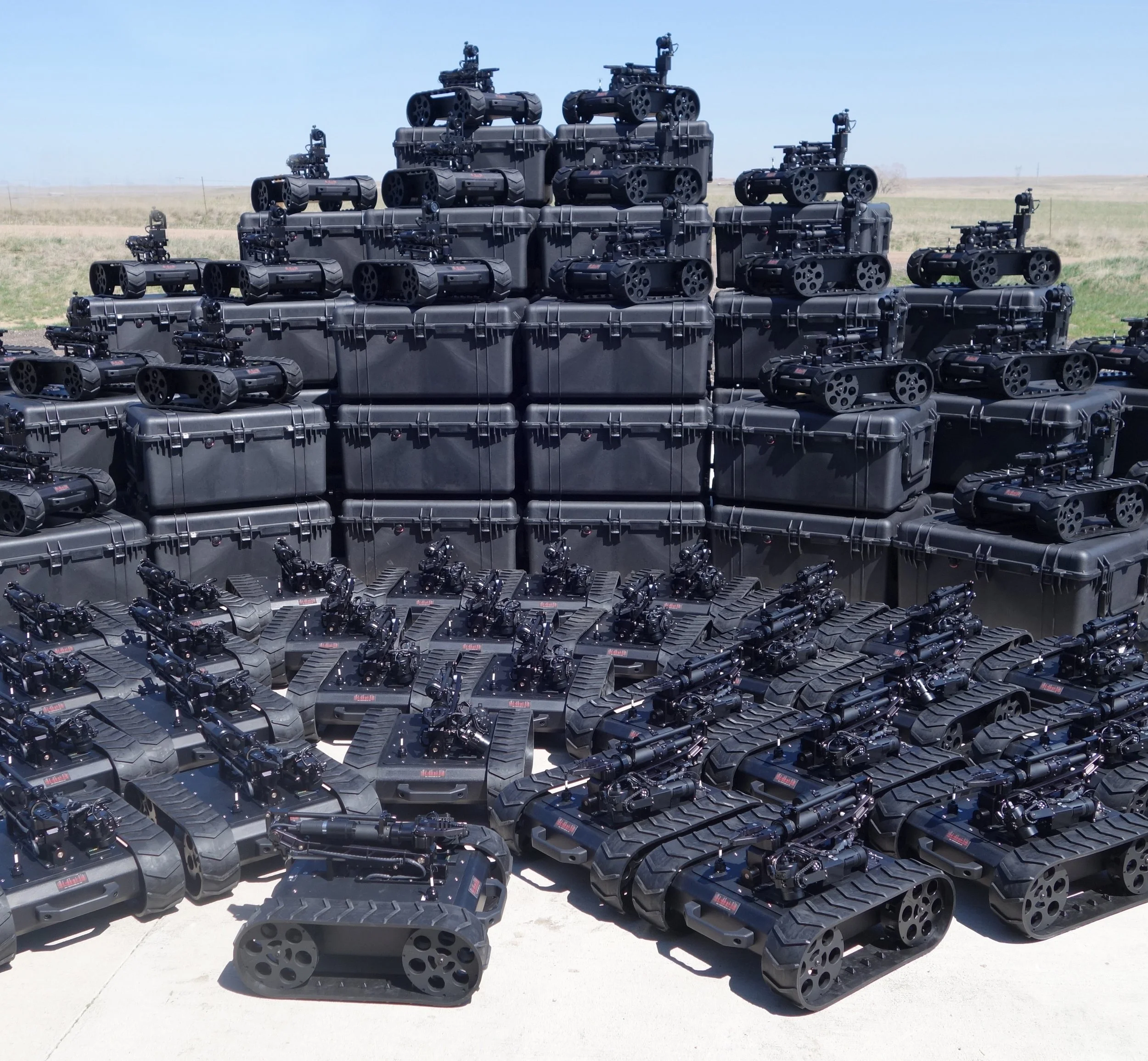 Multiple MMP-30-EOD robots with tracks and cameras stacked outdoors on a paved surface with a grassy field and blue sky in the background.