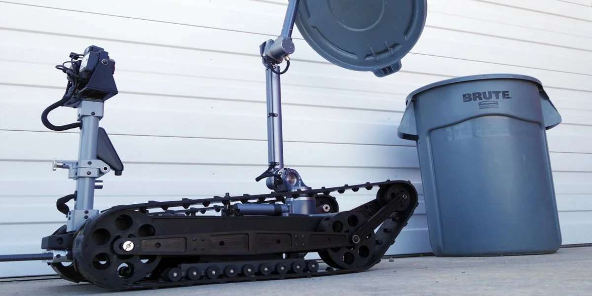 A robotic device with a track system, mounted arm, and a large circular component, placed next to a trash bin against a white siding wall.