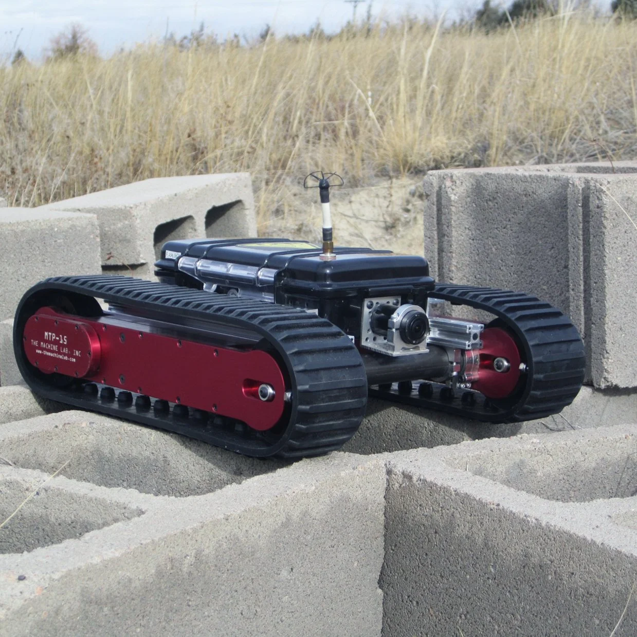 A small robotic tracked vehicle built with our TRACK Pods equipped with a camera and antenna, positioned on concrete blocks outdoors with tall grass in the background.
