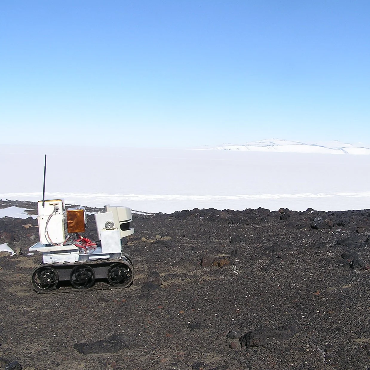 An MMP-40 robot with wheels and antennas on rocky terrain in a cold, snowy landscape under a clear blue sky.