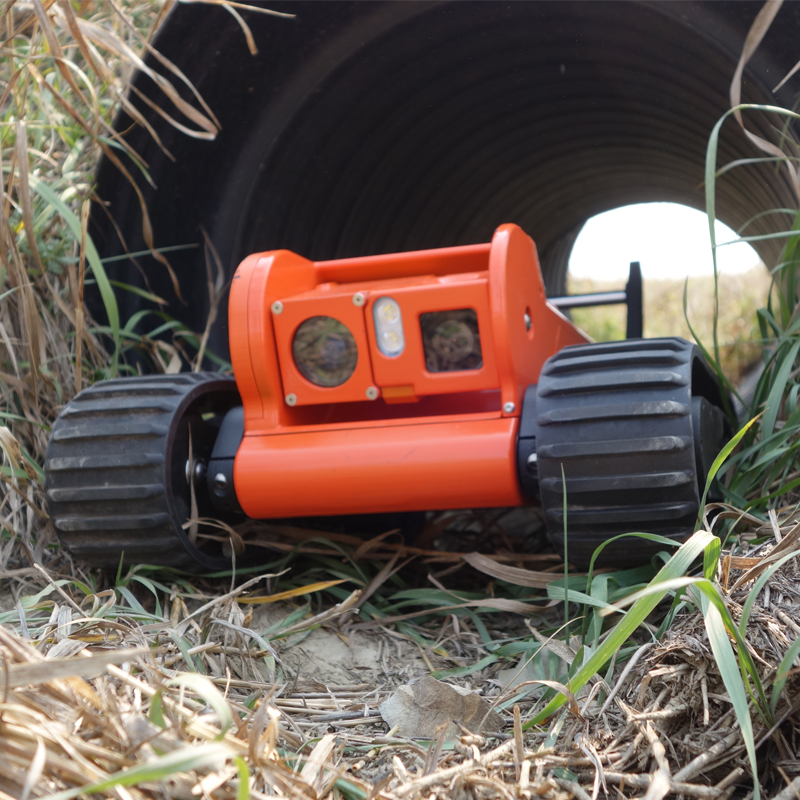 Close-up of the Glimpse inspection robot, remote-controlled with tracks, positioned in a tunnel of foliage and dirt, with a grassy background visible through the opening.
