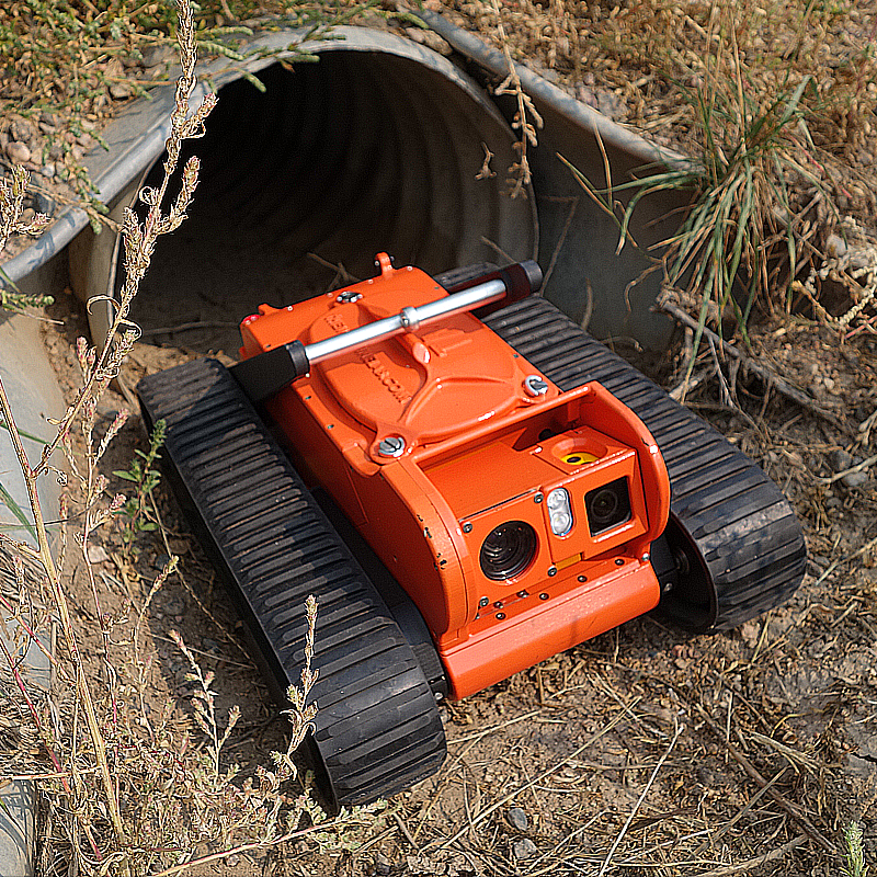 The Glimpse robot with tank-like treads emerging from a drainage pipe in the dirt, surrounded by sparse vegetation.