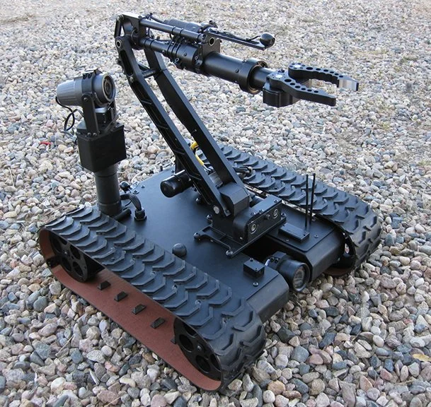 A small robotic vehicle with tank tracks, a mounted camera on a gimbal, and an extendable mechanical arm with a gripping tool at the end, placed on gravel ground.