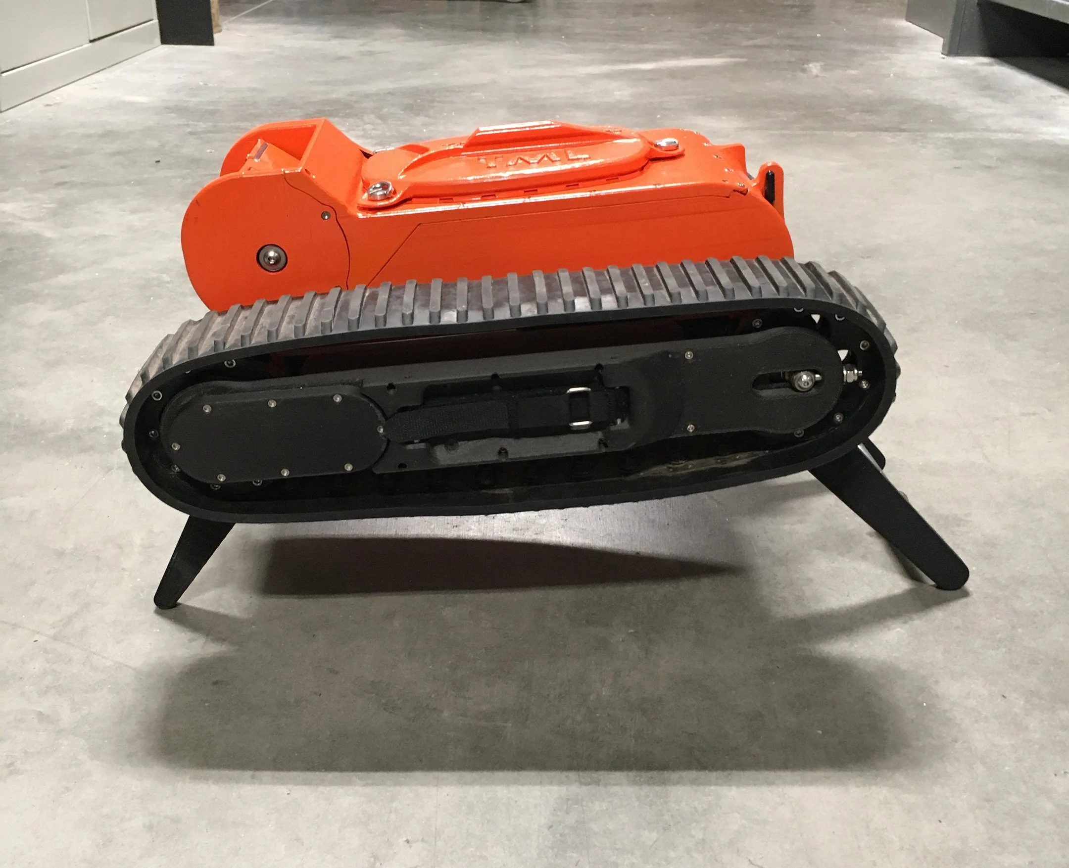 A small, orange remote-controlled robot with tank tracks on a gray floor in a building or warehouse.