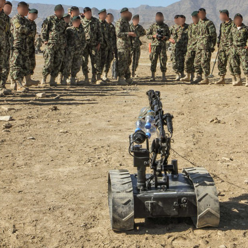 Military personnel gathered outdoors in camouflage uniforms, observing a tracked remote-controlled robotic device equipped with water bottles and sensors.