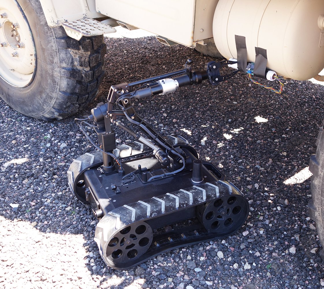 An EOD robot with tracks, inspecting the underside of a large military vehicle, likely used for inspection or maintenance.