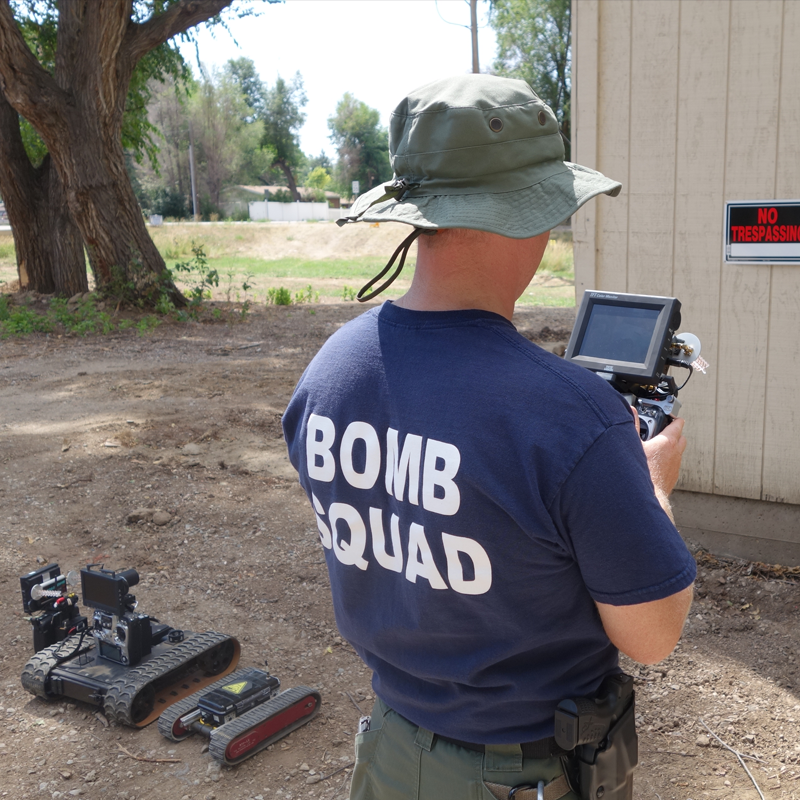 A person wearing a blue t-shirt with 'BOMB SQUAD' on the back, a wide-brim hat, and a holstered gun, operating a remote control device outdoors. There are various remote-controlled devices on the ground nearby, with trees and a building in the backgr