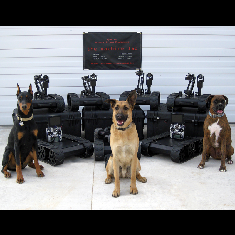 Three dogs sitting in front of four robot tank-like devices with control panels and cameras, inside a warehouse. A sign reads 'the machine lab' with additional descriptions.