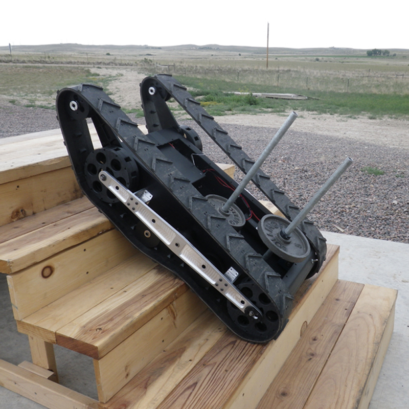 The image shows a shape-changing tracked robot with metal rods protruding from it, sitting on a wooden surface outdoors in a rural area with open fields in the background.