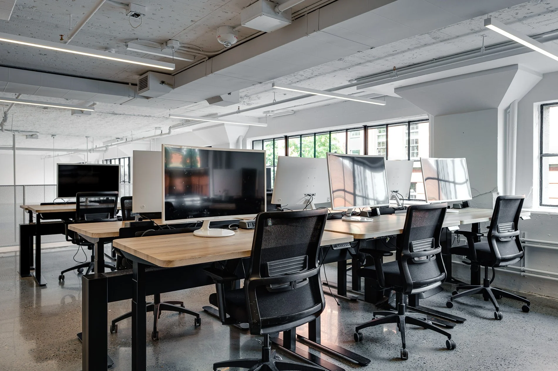 Modern office with desks, black chairs, and multiple large monitors, bright natural light coming through windows, and exposed ceiling with industrial design elements.