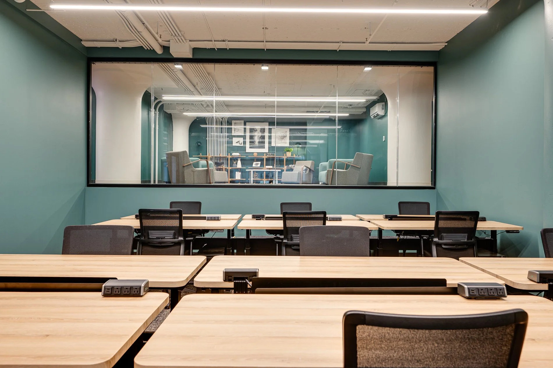 Empty classroom with wooden tables, black chairs, and power outlets, view through a glass partition into a lounge area with armchairs, a bookshelf, and framed pictures.