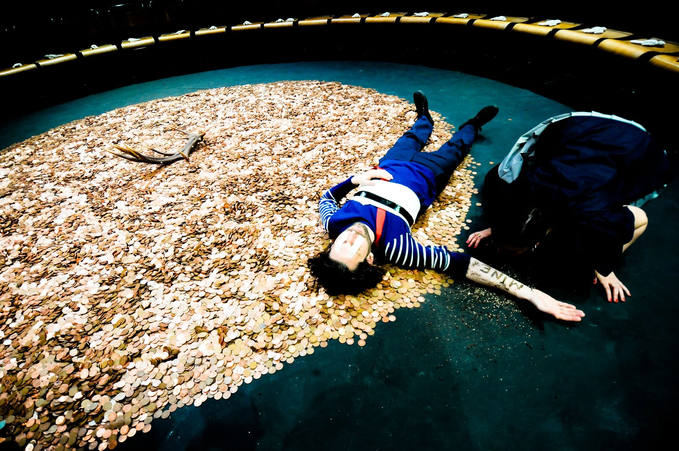 Person dressed as a pirate lying on the ground covered with coins while another person helps them up in a circular enclosure.