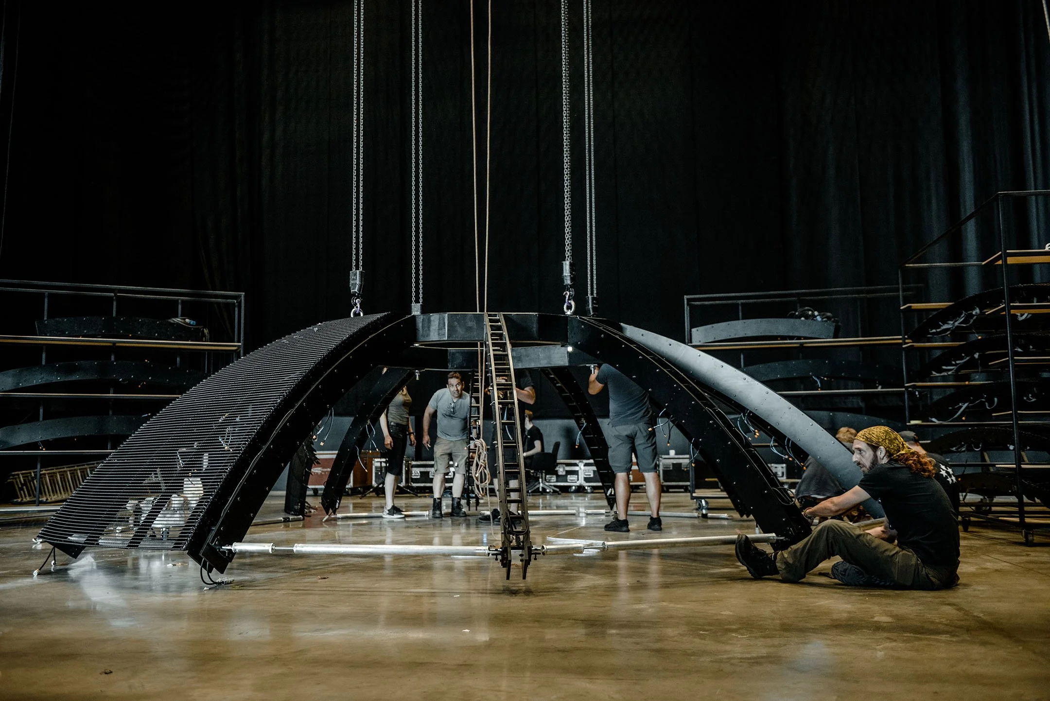People setting up a large black stage or sculpture with curved arches and a ladder, on a wooden floor in a dark theater or auditorium.