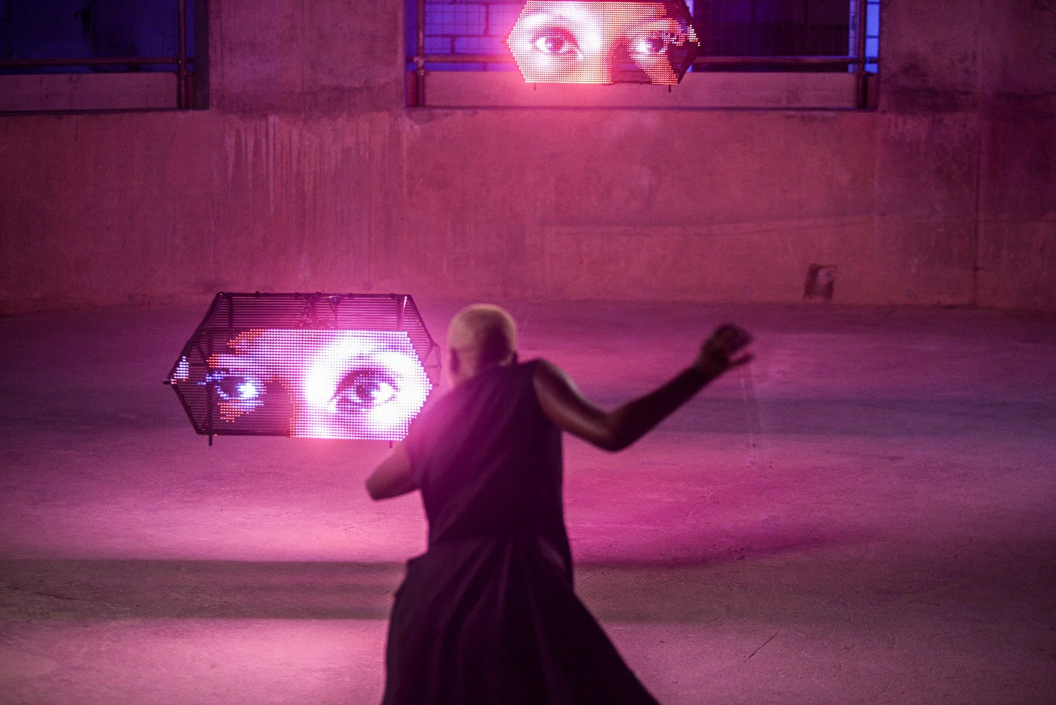 Person with bleached blonde hair and black clothing in front of digital screens displaying a pair of eyes, in a pink-lit, industrial space.