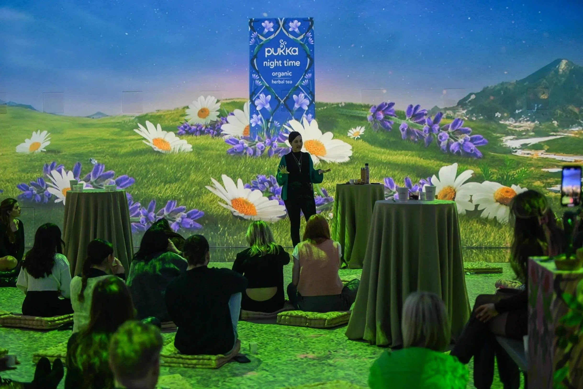A group of people seated on mats facing a woman giving a presentation, with a large digital backdrop showing a field of daisies and lavender, and a sign that reads 'pukka nighttime organic herbal tea'.