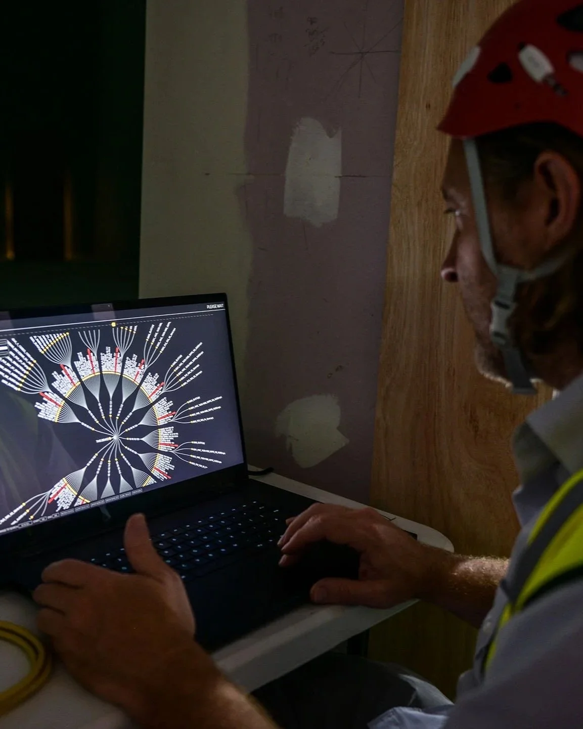 A man wearing a red helmet working on a laptop in a dimly lit room, with a diagram or chart displayed on the screen.
